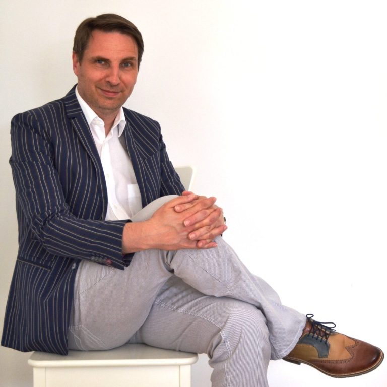 Der Autor sitzt mit verschränkten Händen und übergeschlagenen Beinen auf einem weißen Stuhl und  trägt ein gestreiftes Sakko.