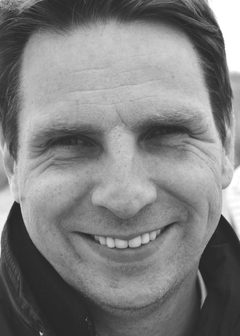 Schwarz-weiß-Porträt eines Mannes mit kurzem Haar und dunkler Jacke vor unscharfem Küsten-Hintergrund.