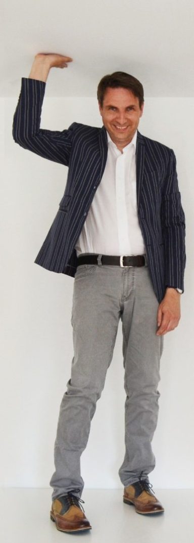 Ein Mann in gestreiftem Sakko, weißem Hemd und grauer Hose steht vor einer weißen Wand.