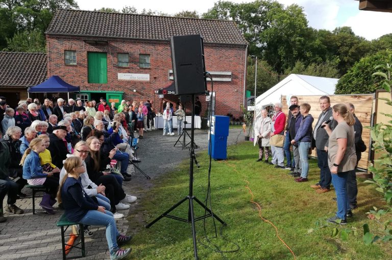 Mehrere Personen stehen nebeneinander auf einer Wiese vor einem Publikum, das auf Bänken vor einem Backsteingebäude sitzt.