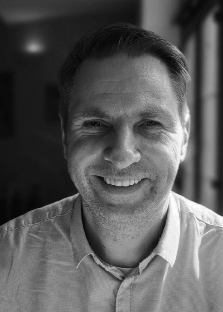 Mann mit kurz geschnittenem Haar trägt ein helles Hemd mit offenem Kragen in einem Innenraum mit Fenster im Hintergrund.