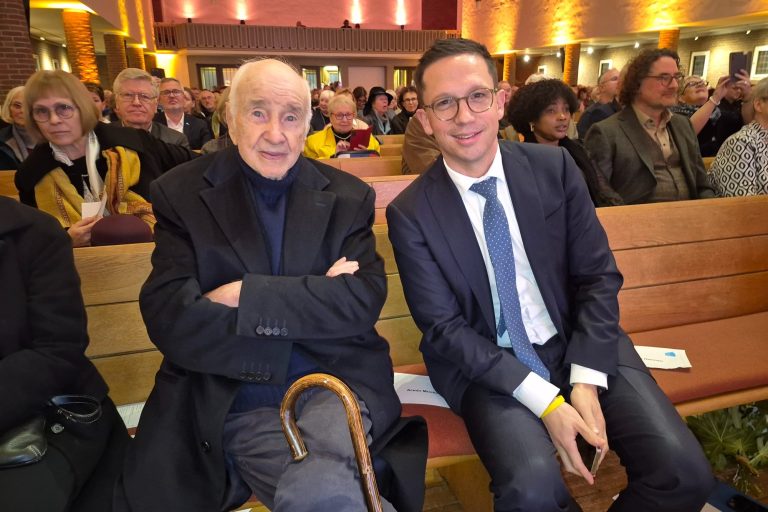 2 Männer in Anzug und Mantel sitzen in einer Kirchenbank, der eine hält einen Gehstock, im Hintergrund Publikum in einer beleuchten Kirche.